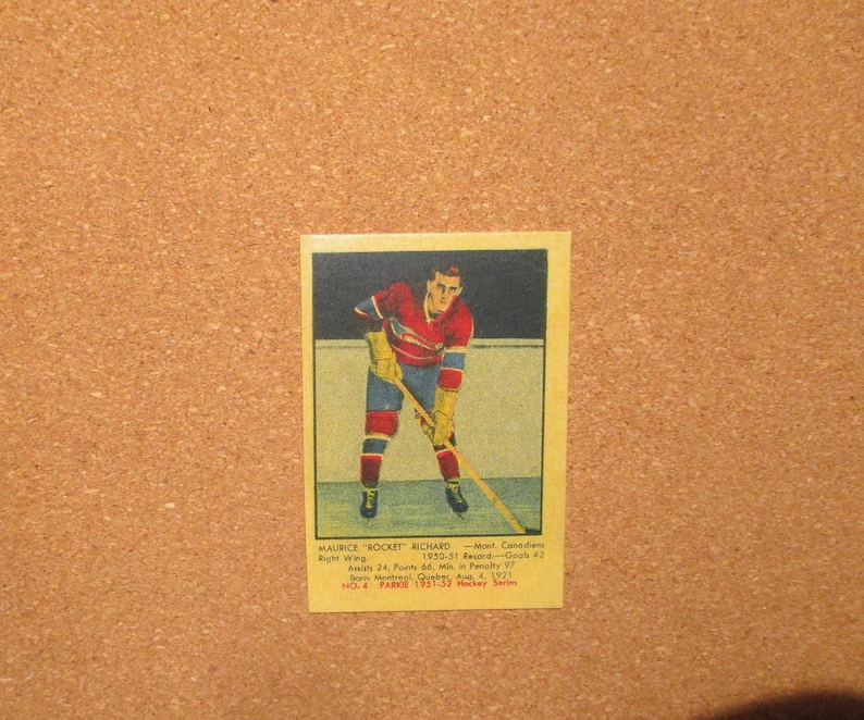 May include: A vintage rectangular hockey trading card featuring an illustration of Maurice Rocket Richard in a red and blue uniform holding a hockey stick. The card includes printed text detailing his 1950-51 record, position, and birth date.