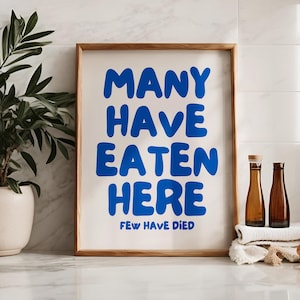 Puede incluir: Un cartel enmarcado con el texto "MANY HAVE EATEN HERE FEW HAVE DIED" en letras azules. El cartel est&aacute; en un marco de madera y sobre una superficie blanca. Hay una planta en maceta a la izquierda y dos botellas a la derecha.