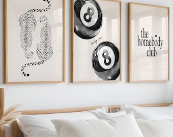 May include: Three framed prints on a white wall above a white bed with a beige throw blanket. The prints are black and white and feature a cheetah, a pool ball, and the text "the homebody club".