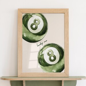 May include: A framed print featuring two green watercolor-style eight balls. The balls have a white eight in the center. The words "lucky me" are written in cursive. The frame is light brown and sits on a green shelf.