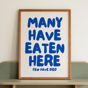 May include: A framed print with the text "MANY HAVE EATEN HERE FEW HAVE DIED" in blue on a white background. The frame is wood-toned, and the print rests on a sage green shelf with rounded edges. The shelf is mounted on a white wall.