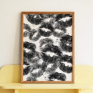 May include: A framed artwork featuring a pattern of black lipstick kiss marks on a white background. The artwork is displayed on a yellow shelf against a white wall. The frame is made of wood.