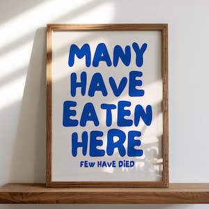 Puede incluir: Un cartel enmarcado con el texto "MANY HAVE EATEN HERE FEW HAVE DIED" en azul sobre un fondo blanco. El marco es de color marr&oacute;n c&aacute;lido y el cartel se exhibe en una estanter&iacute;a de madera.
