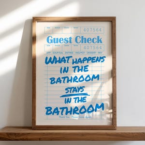 May include: A framed print with the words "Guest Check" and "What happens in the bathroom stays in the bathroom" in blue text. The print is in a wooden frame and rests on a wooden shelf against a white wall.