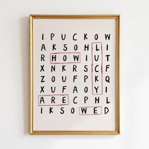 May include: A framed wall art print featuring a word search puzzle on a white background with black letters. The words HOW, LUCKY, ARE, and WED are highlighted with red rectangular outlines. The frame is a thin, metallic gold color.