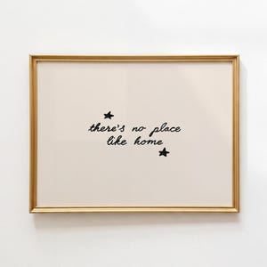 Pu&ograve; includere: Una stampa artistica rettangolare in una sottile cornice dorata con sfondo color crema e il testo nero scritto a mano there's no place like home centrato sulla pagina, accompagnato da due piccole stelle nere disegnate a mano sopra e sotto.