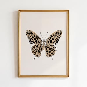 May include: A framed wall art print featuring a centered butterfly illustration with a tan background and black leopard print spots on the wings. The butterfly is symmetrical with thin antennae and is enclosed in a thin, metallic gold frame.