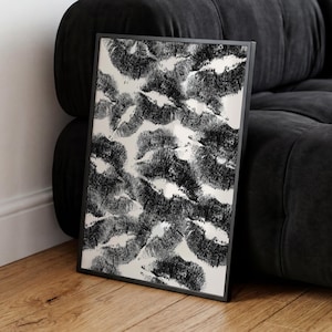 May include: Black and white framed art print featuring a pattern of overlapping lip imprints. The artwork is set against a light background and framed in a black border. The print is leaning against a black sofa on a wooden floor.