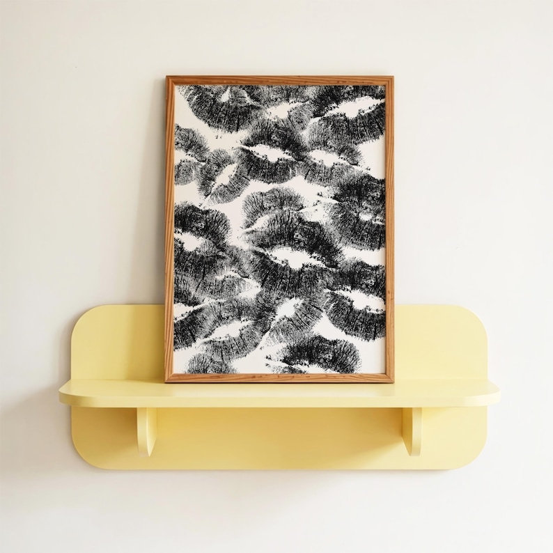 May include: A framed artwork featuring a pattern of black lipstick kiss marks on a white background. The artwork is displayed on a yellow shelf against a white wall. The frame is made of wood.