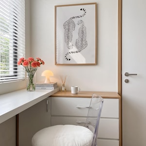 May include: A framed wall art print featuring two black and white cheetah illustrations, displayed above a white desk area with a clear chair, a glass vase of pink carnations, a small mushroom-shaped lamp, and a white ceramic coffee mug.