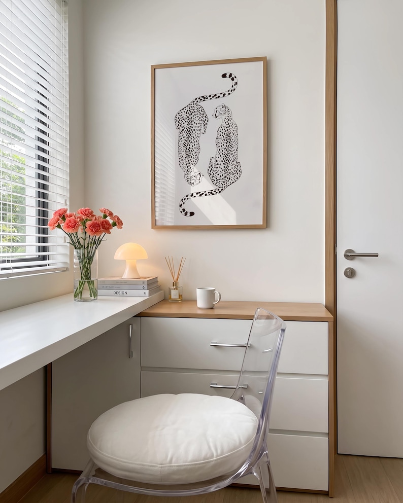 May include: A framed wall art print featuring two black and white cheetah illustrations, displayed above a white desk area with a clear chair, a glass vase of pink carnations, a small mushroom-shaped lamp, and a white ceramic coffee mug.