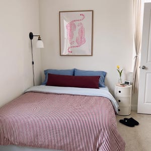 May include: A bed with a light blue sheet and a burgundy and white striped comforter. A long burgundy bolster pillow sits in front of two blue pillows. Above the bed is a framed print of two pink leopards. A wall lamp and a round nightstand are present.