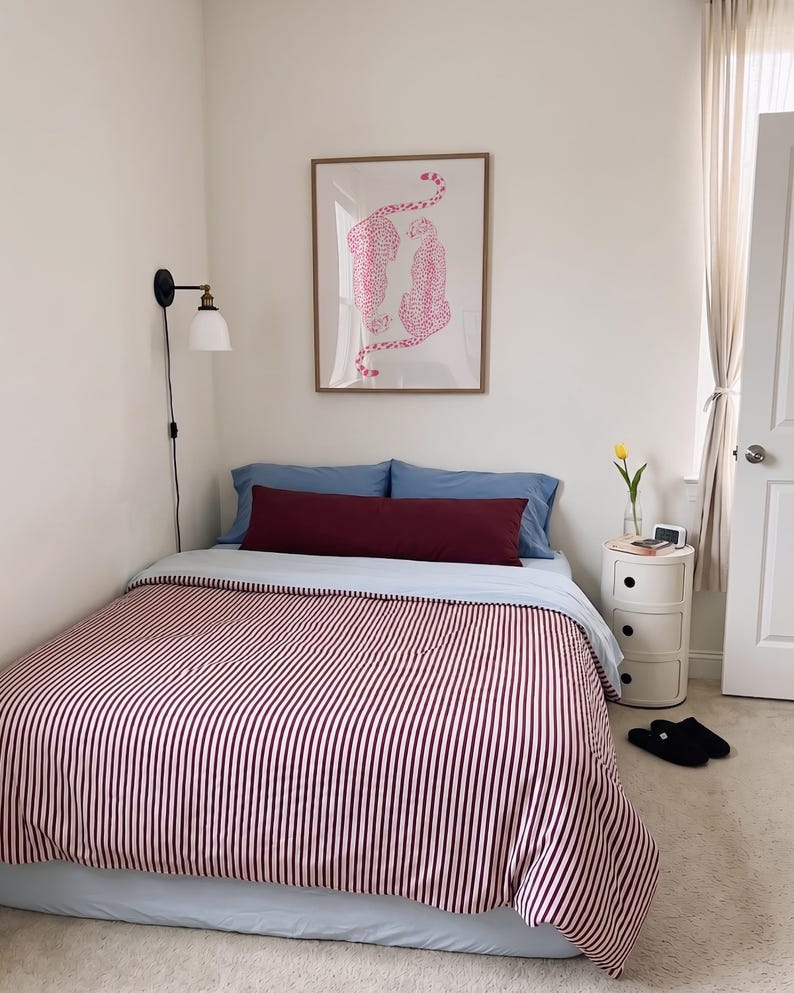 May include: A bed with a light blue sheet and a burgundy and white striped comforter. A long burgundy bolster pillow sits in front of two blue pillows. Above the bed is a framed print of two pink leopards. A wall lamp and a round nightstand are present.