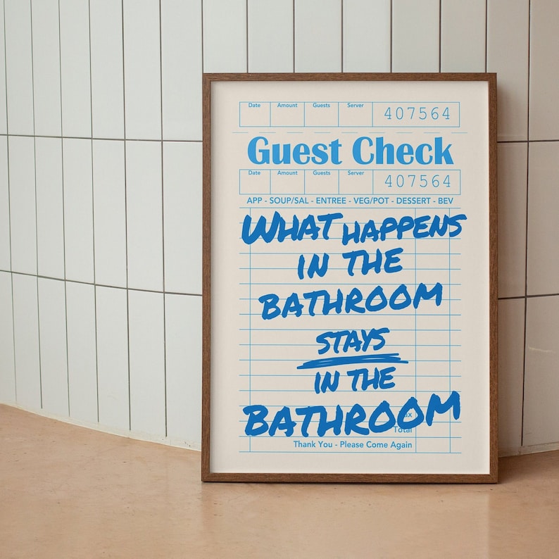 May include: A framed print with a brown wooden frame. The print has a light beige background with blue lines and text that reads "Guest Check" and "What happens in the bathroom stays in the bathroom."