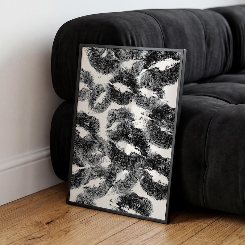 May include: Black and white framed art print featuring a pattern of overlapping lip imprints. The artwork is set against a light background and framed in a black border. The print is leaning against a black sofa on a wooden floor.