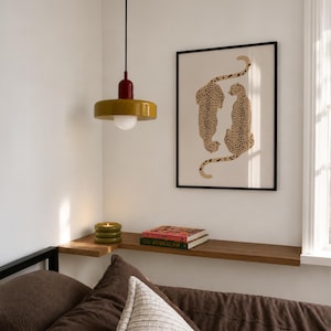 May include: A mustard yellow pendant light with a red top and white bulb, a framed print featuring two spotted cheetahs, a green stacked candle, and two books on a wooden shelf above a brown bed with a textured white pillow.