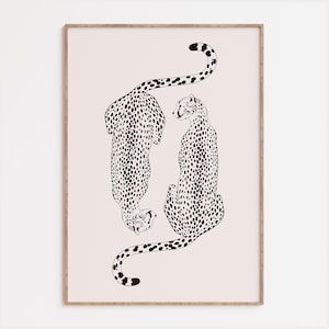 May include: A framed wall art print featuring a black line drawing of two cheetahs on a pale pink background. One cheetah is positioned facing downward while the other faces upward, both covered in black spots, enclosed in a light wood frame.