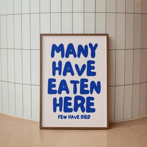 Puede incluir: Un p&oacute;ster enmarcado con las palabras "MANY HAVE EATEN HERE" en letras azules en negrita sobre un fondo blanco. Debajo, en texto azul m&aacute;s peque&ntilde;o, se lee "FEW HAVE DIED". El marco es de color marr&oacute;n.