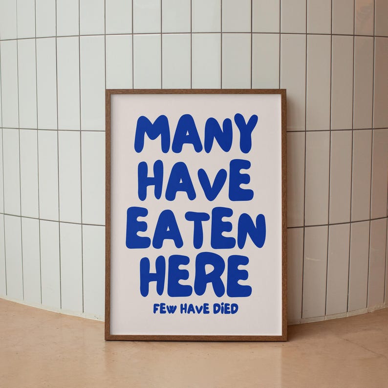 Puede incluir: Un p&oacute;ster enmarcado con las palabras "MANY HAVE EATEN HERE" en letras azules en negrita sobre un fondo blanco. Debajo, en texto azul m&aacute;s peque&ntilde;o, se lee "FEW HAVE DIED". El marco es de color marr&oacute;n.