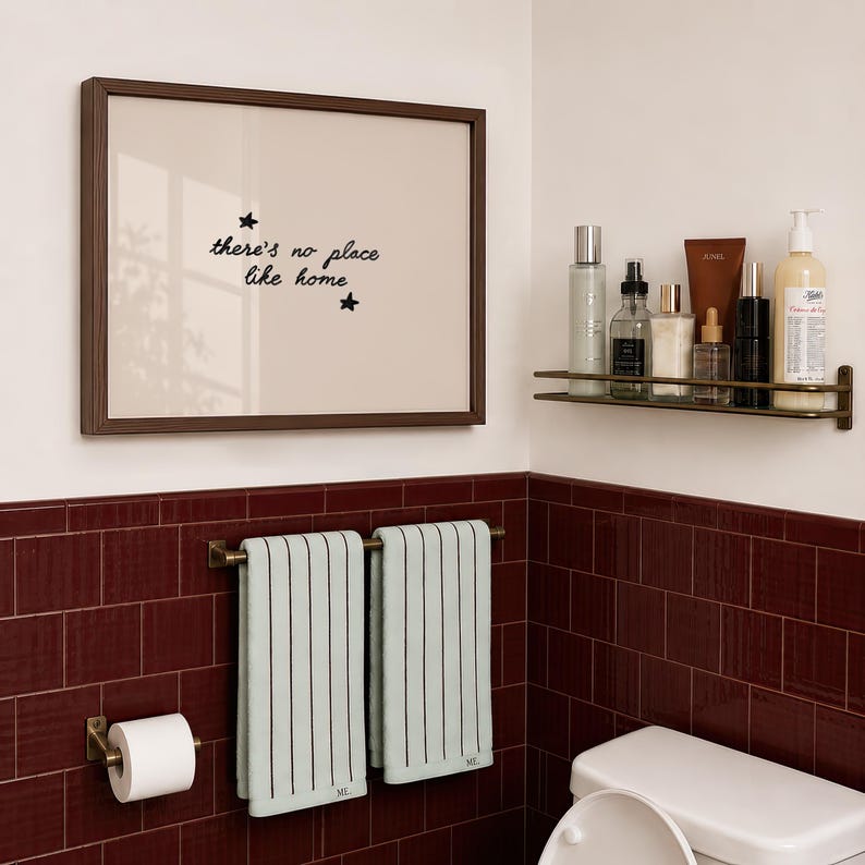 Pu&ograve; includere: Una cornice rettangolare da parete con bordo marrone scuro contenente il testo "there's no place like home" con due piccole stelle. Sotto, una mensola in metallo sostiene vari flaconi e un portasciugamani espone due asciugamani a righe verde chiaro.