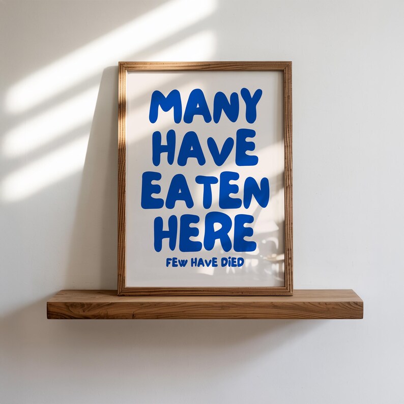Puede incluir: Un cartel enmarcado con el texto "MANY HAVE EATEN HERE FEW HAVE DIED" en azul sobre un fondo blanco. El marco es de color marr&oacute;n c&aacute;lido y el cartel se exhibe en una estanter&iacute;a de madera.