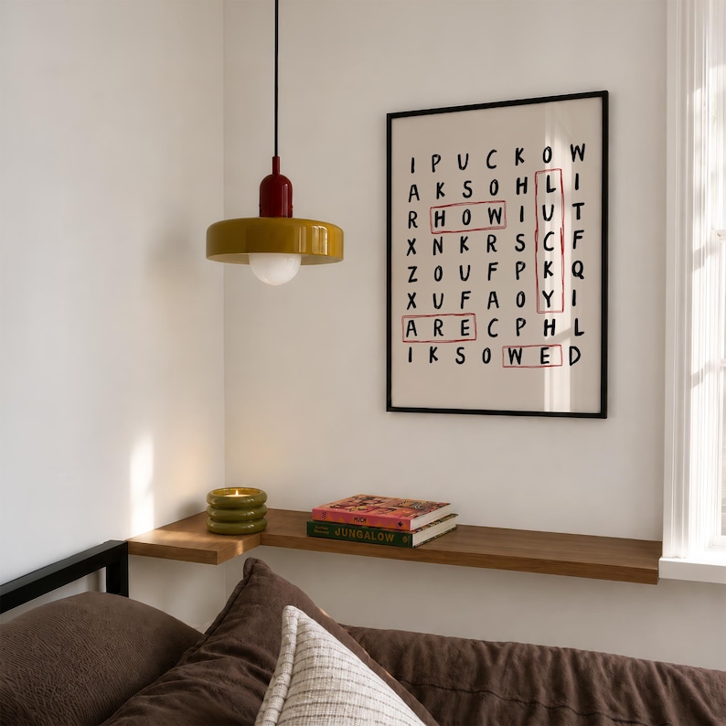 May include: A mustard yellow pendant light with a red top and white bulb hangs above a wooden shelf. On the wall, a framed word search print features the circled words HOW, LUCKY, ARE, and WED in red ink against a cream background.