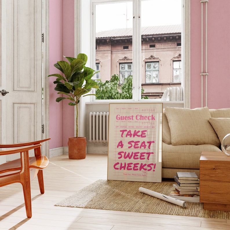 K&ouml;nnte beinhalten: Ein rosa-wei&szlig;es G&auml;ste-Check-Schild mit dem Text "Take a seat sweet cheeks!" in rosa Schrift. Das Schild ist in Gold gerahmt und lehnt an einem beigen Sofa in einem Raum mit einer rosa Wand und einem gro&szlig;en Fenster.