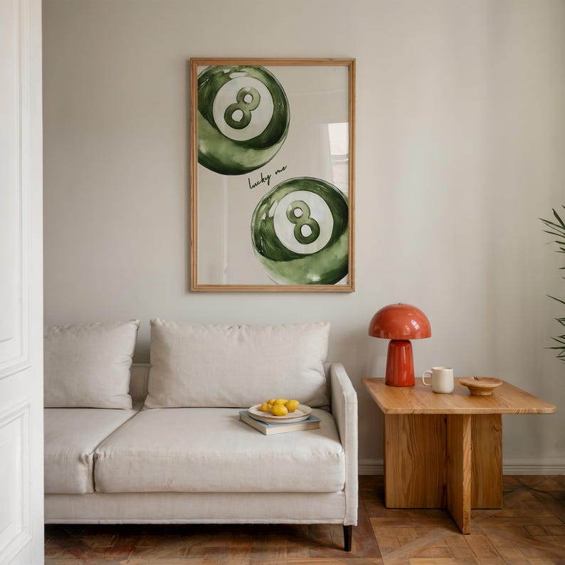 May include: A framed print featuring two green and white watercolor-style billiard balls with the number eight. The words "lucky me" are written below the top ball. The print hangs above a cream-colored sofa.
