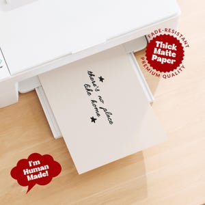 Pu&ograve; includere: Una stampante bianca che espelle un foglio di carta opaca spessa con il testo stampato in nero there's no place like home e due piccole stelle. Un badge circolare rosso riporta Fade-Resistant Premium Quality e un fumetto rosso dice I'm Human Made.