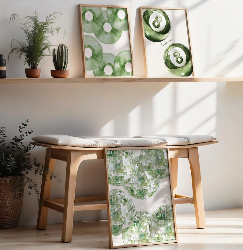 May include: Three framed art prints with watercolor designs. One features green vinyl records, another shows a green eight ball, and the third displays green disco balls. The prints are displayed on a wooden shelf and bench, with potted plants.