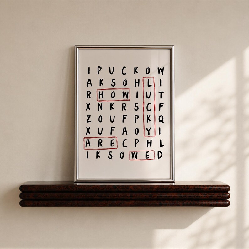 May include: A framed wall art print featuring a word search puzzle on a white background with black letters. The words HOW, LUCKY, ARE, and WED are highlighted with red rectangular outlines. The print is enclosed in a thin silver metal frame.