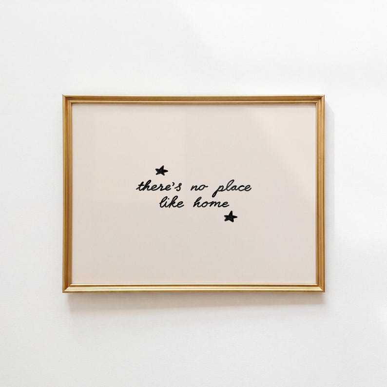 Pu&ograve; includere: Una stampa artistica rettangolare in una sottile cornice dorata con sfondo color crema e il testo nero scritto a mano there's no place like home centrato sulla pagina, accompagnato da due piccole stelle nere disegnate a mano sopra e sotto.