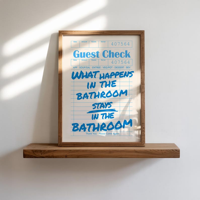 May include: A framed print with the words "Guest Check" and "What happens in the bathroom stays in the bathroom" in blue text. The print is in a wooden frame and rests on a wooden shelf against a white wall.