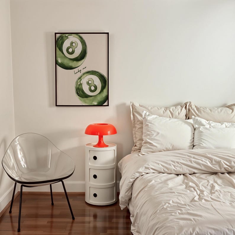 May include: A bedroom scene featuring a framed print with two green eight-ball illustrations and the text "lucky me." An orange mushroom-shaped lamp sits atop a white bedside table with three drawers. A clear plastic chair and a bed with beige bedding complete the room.