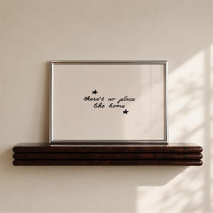 Pu&ograve; includere: Una cornice rettangolare argentata contenente una stampa beige chiaro con il testo nero scritto a mano there's no place like home, accentuato da due piccole stelle nere disegnate a mano sopra e sotto la frase centrata.