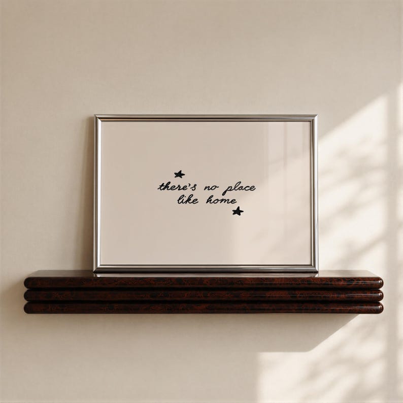 Pu&ograve; includere: Una cornice rettangolare argentata contenente una stampa beige chiaro con il testo nero scritto a mano there's no place like home, accentuato da due piccole stelle nere disegnate a mano sopra e sotto la frase centrata.