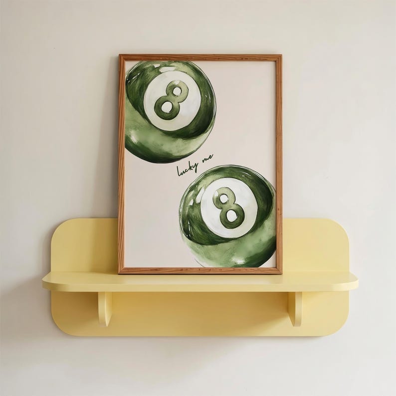 May include: A framed print featuring two green watercolor-style eight balls with white number 8s. The words "lucky me" are written in cursive. The print is displayed on a yellow shelf.