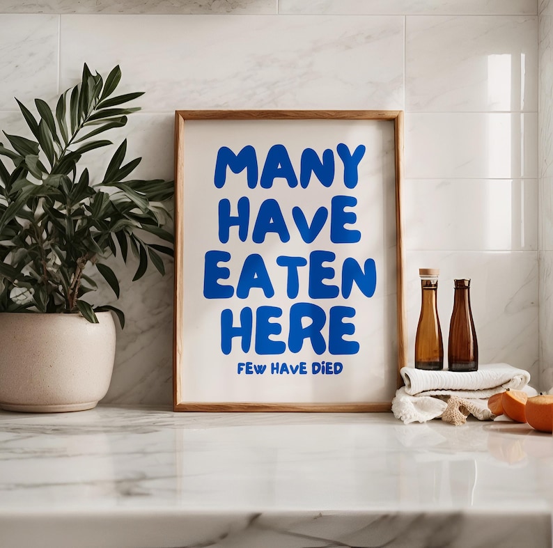 Puede incluir: Un cartel enmarcado con el texto "MANY HAVE EATEN HERE FEW HAVE DIED" en letras azules. El cartel est&aacute; en un marco de madera y sobre una superficie blanca. Hay una planta en maceta a la izquierda y dos botellas a la derecha.