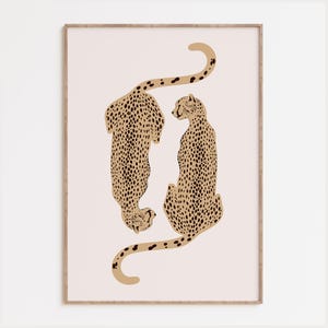 May include: A framed wall art print featuring two stylized cheetahs in tan with black spots, positioned vertically in a mirrored orientation against a pale pink background. The cheetahs are depicted with long, curved tails and detailed markings.