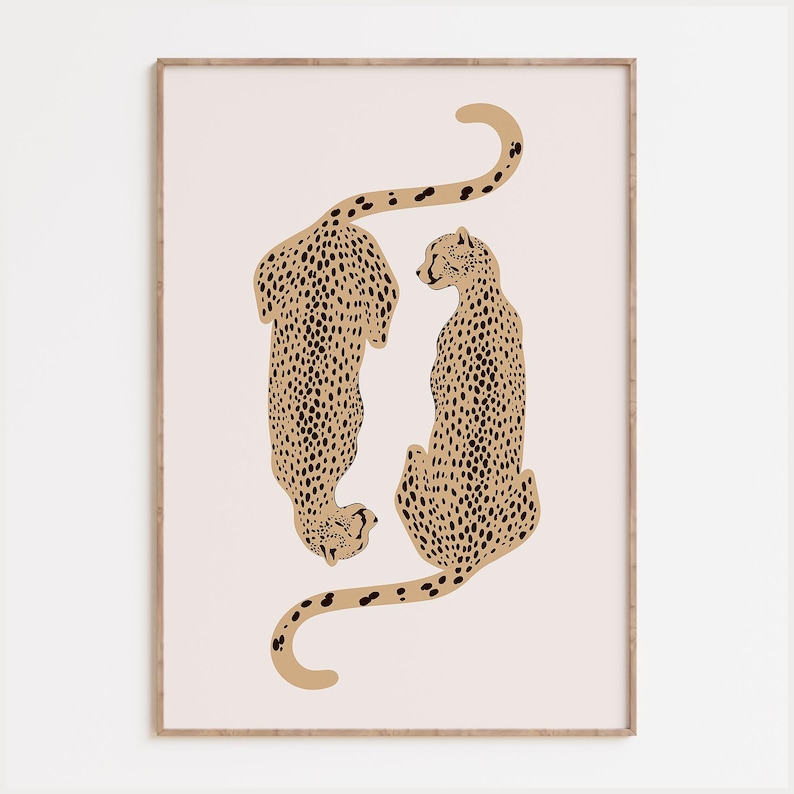 May include: A framed wall art print featuring two stylized cheetahs in tan with black spots, positioned vertically in a mirrored orientation against a pale pink background. The cheetahs are depicted with long, curved tails and detailed markings.