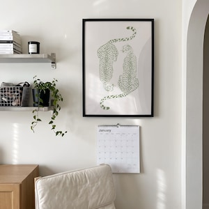 May include: A framed wall art print featuring two green cheetahs with spotted patterns on a light background, positioned above a white wall calendar with the text January printed at the top, displayed on a plain white wall in a room.