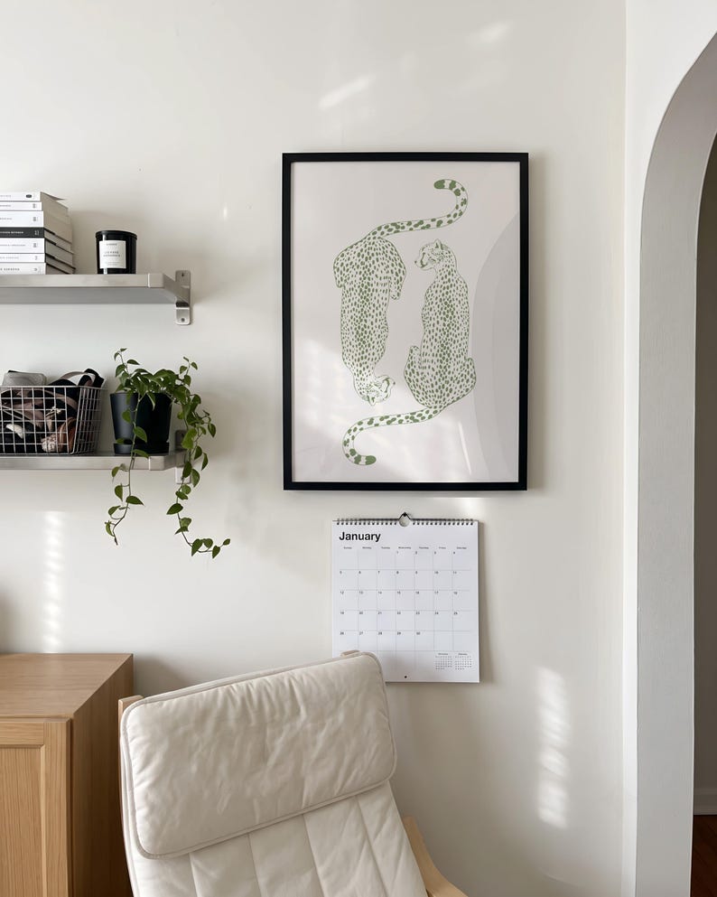 May include: A framed wall art print featuring two green cheetahs with spotted patterns on a light background, positioned above a white wall calendar with the text January printed at the top, displayed on a plain white wall in a room.