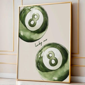 May include: A framed print featuring two green and white eight-ball pool balls. The balls have a watercolor effect and are set against a neutral background. The words "lucky me" are written in cursive below the top ball.