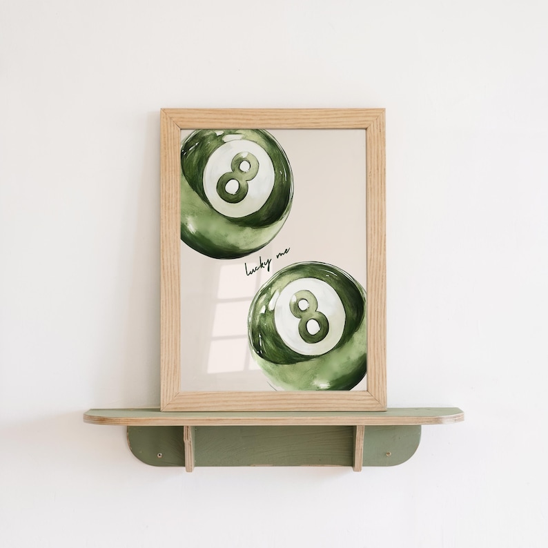 May include: A framed print featuring two green watercolor-style eight balls. The balls have a white eight in the center. The words "lucky me" are written in cursive. The frame is light brown and sits on a green shelf.