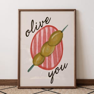 May include: A framed wall art print featuring a watercolour illustration of three green olives on a toothpick, centred over a red and pink striped circle. The black cursive text reads olive above the olives and you below the olives.