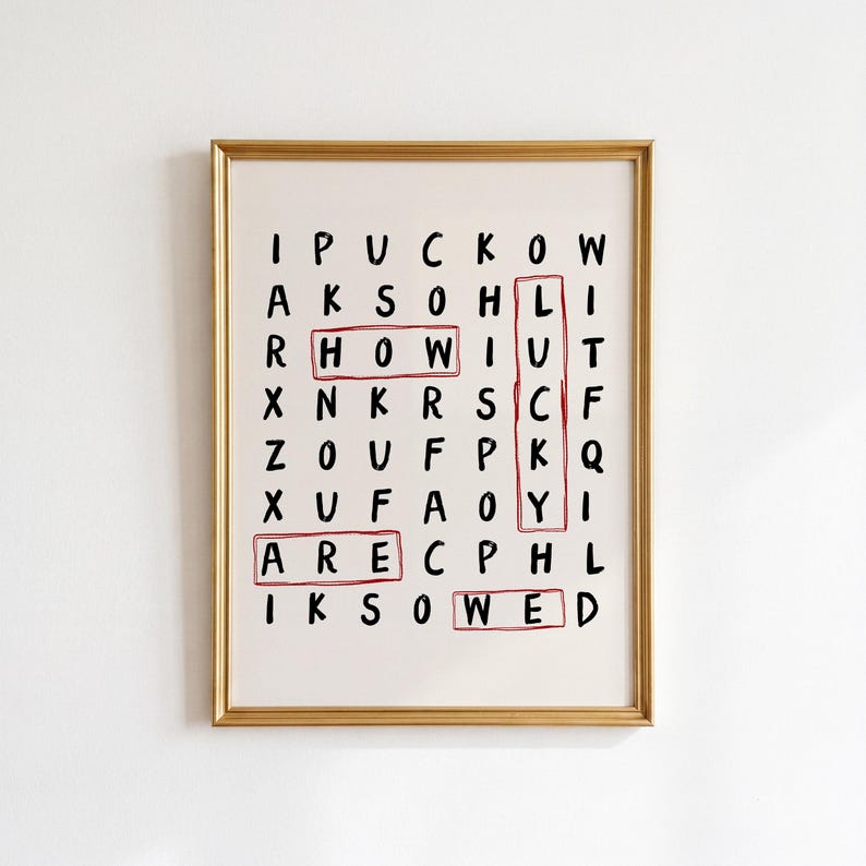May include: A framed wall art print featuring a word search puzzle on a white background with black letters. The words HOW, LUCKY, ARE, and WED are highlighted with red rectangular outlines. The frame is a thin, metallic gold color.