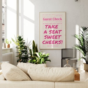 K&ouml;nnte beinhalten: Ein rosa-wei&szlig;er Gastcheck mit dem Text "Take a seat sweet cheeks!" darauf gedruckt. Der Check ist gerahmt und h&auml;ngt an einer wei&szlig;en Wand.