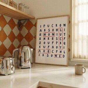 May include: A framed word search print with black letters on a white background. The words HOW, ARE, WED, and LUCKY are highlighted with red rectangular outlines. The print is displayed on a kitchen counter next to a silver kettle and a mug.