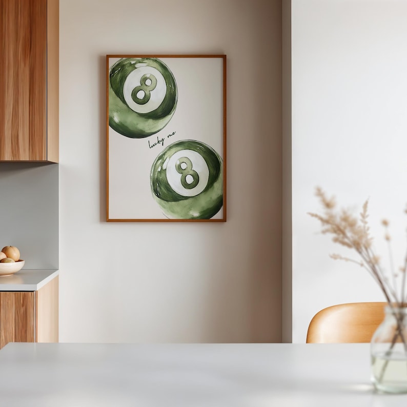 May include: A framed print featuring two green and white watercolor-style eight-ball illustrations. The phrase "lucky me" is written in cursive below the top ball. The frame is a warm brown color, and the print hangs on a neutral wall.