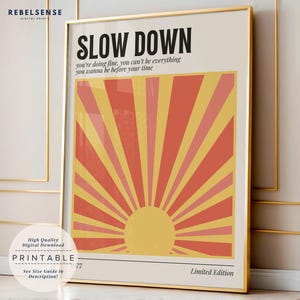 Peut inclure: Une affiche imprimable avec un soleil jaune se levant sur un fond rayé rouge et rose. Le texte "SLOW DOWN" est en haut de l'affiche, et le texte "you're doing fine, you can't be everything you wanna be before your time" est sous le soleil. L'affiche indique également "Édition limitée".